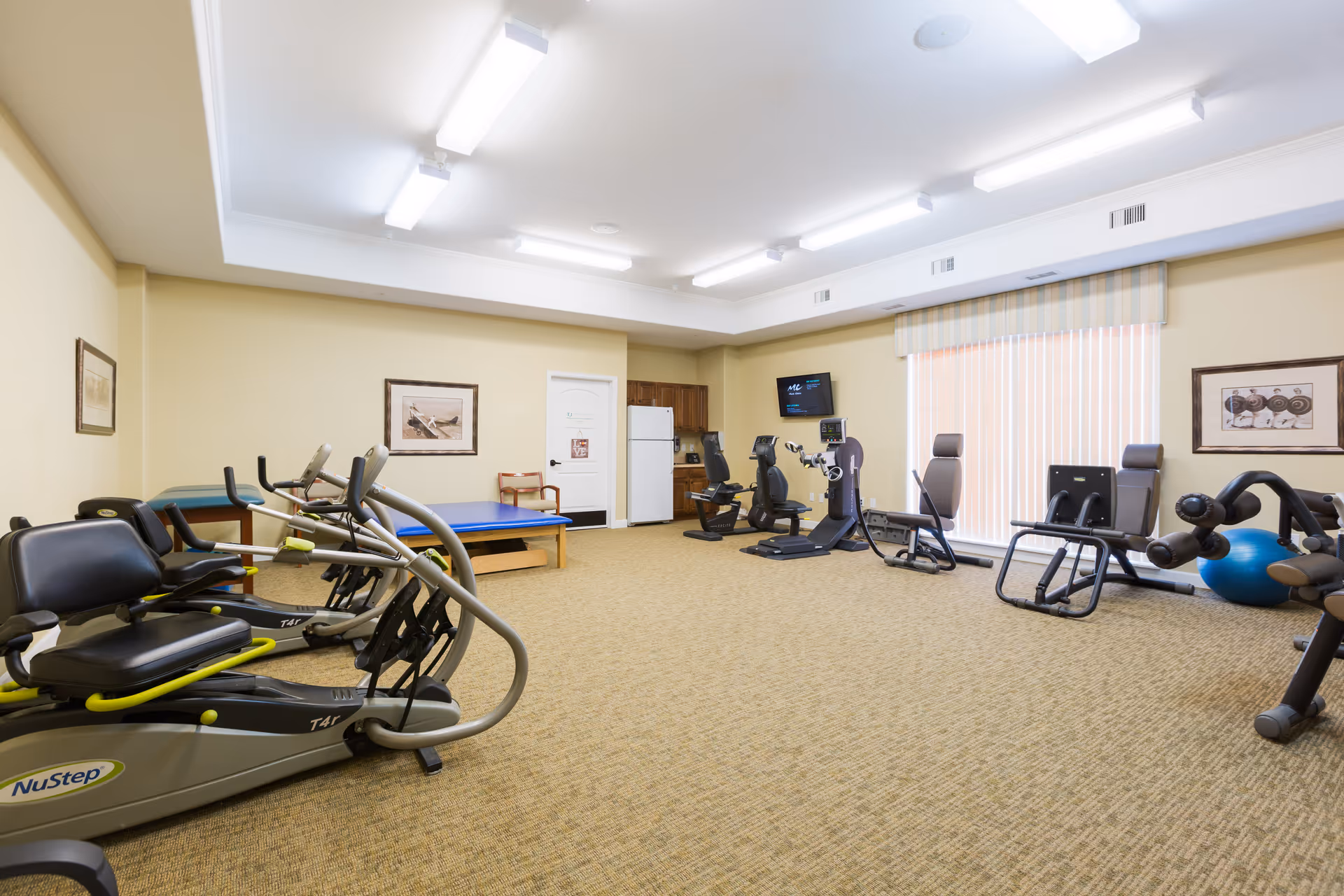 A spacious fitness room in an assisted living facility featuring several exercise machines including recumbent bikes, a rowing machine, and other workout equipment. The room has beige walls, carpeted flooring, fluorescent ceiling lights, framed pictures on the walls, a television mounted on the far wall, and a window with vertical blinds.