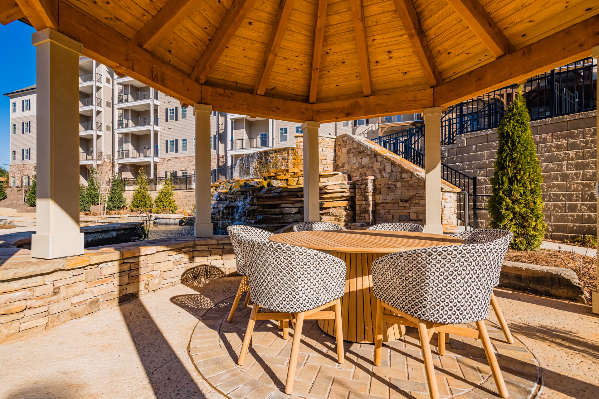 Outdoor seating area under a wooden gazebo with a round table and four patterned chairs. In the background, there is a stone waterfall feature, landscaping with small trees, and a multi-story residential building.