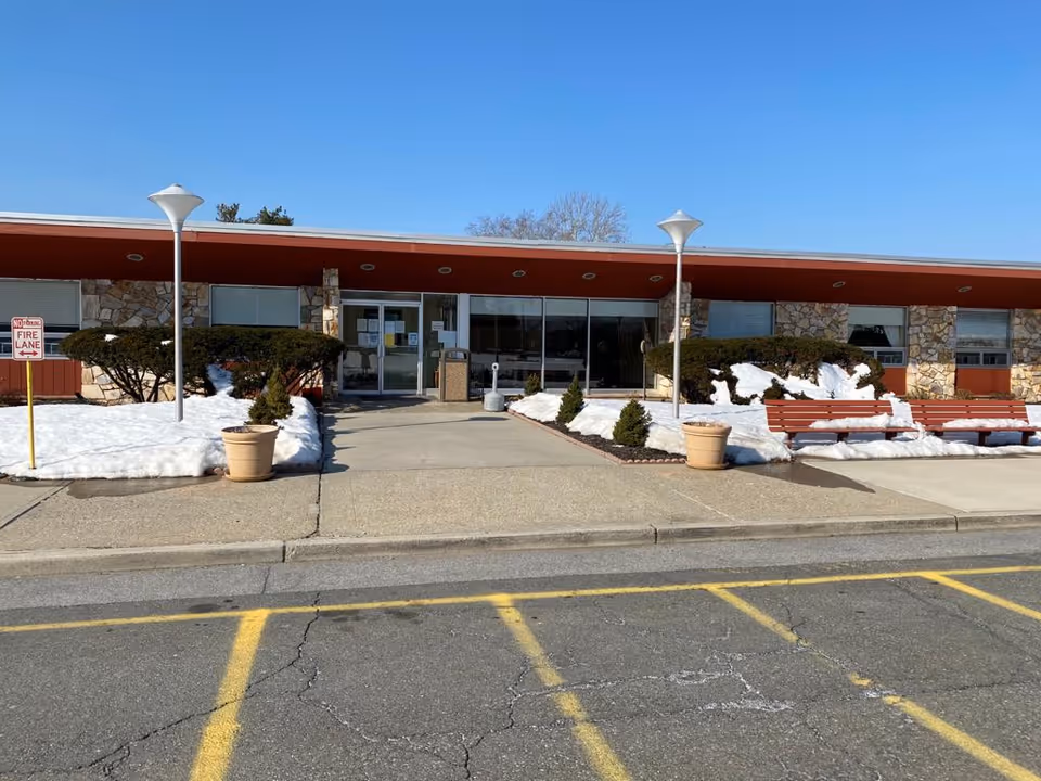 Front exterior view of Limecrest Subacute and Rehabilitation Center building with a flat roof, stone and red panel facade, glass entrance doors, snow-covered bushes, two lampposts, two large potted plants, and two red benches on the right side. The parking area with yellow lines is visible in the foreground under a clear blue sky.