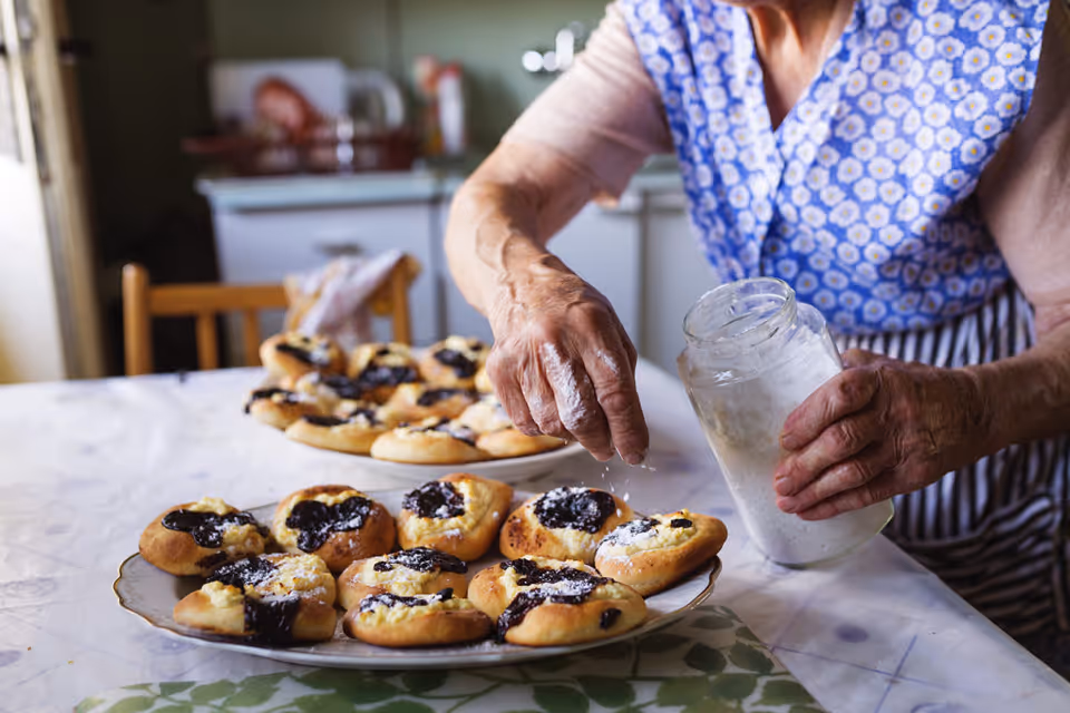 A person dusting powdered sugar onto freshly baked pastries arranged on plates in a home kitchen.
