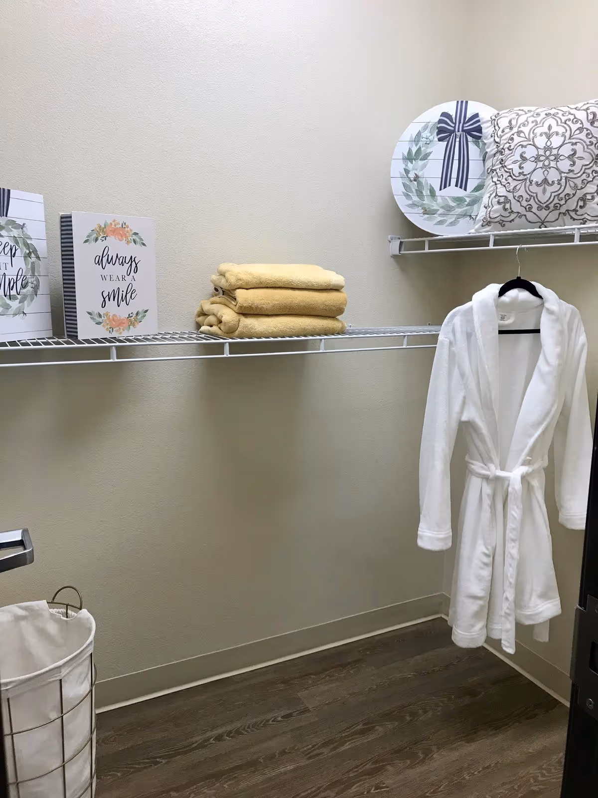 A small room with beige walls and wood flooring featuring a white wire shelf holding folded yellow towels, decorative signs, a round decorative plate, and a patterned pillow. A white bathrobe hangs on a hanger from the shelf, and a laundry basket with a white liner is on the floor.