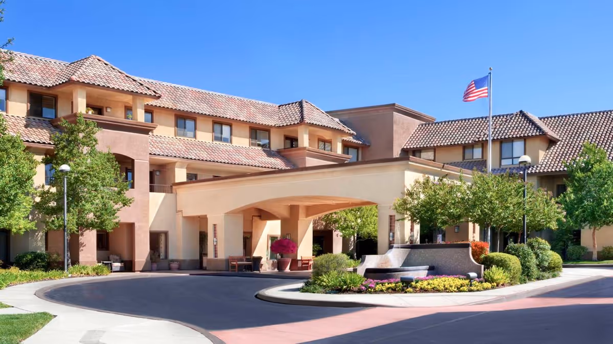 Exterior view of Ivy Park at Roseville, a multi-story senior living facility with beige and brown stucco walls and red tile roofing. The entrance features a covered drop-off area with benches and potted plants. There is a landscaped roundabout with flowers and shrubs, and an American flag flying on a flagpole. The sky is clear and blue.