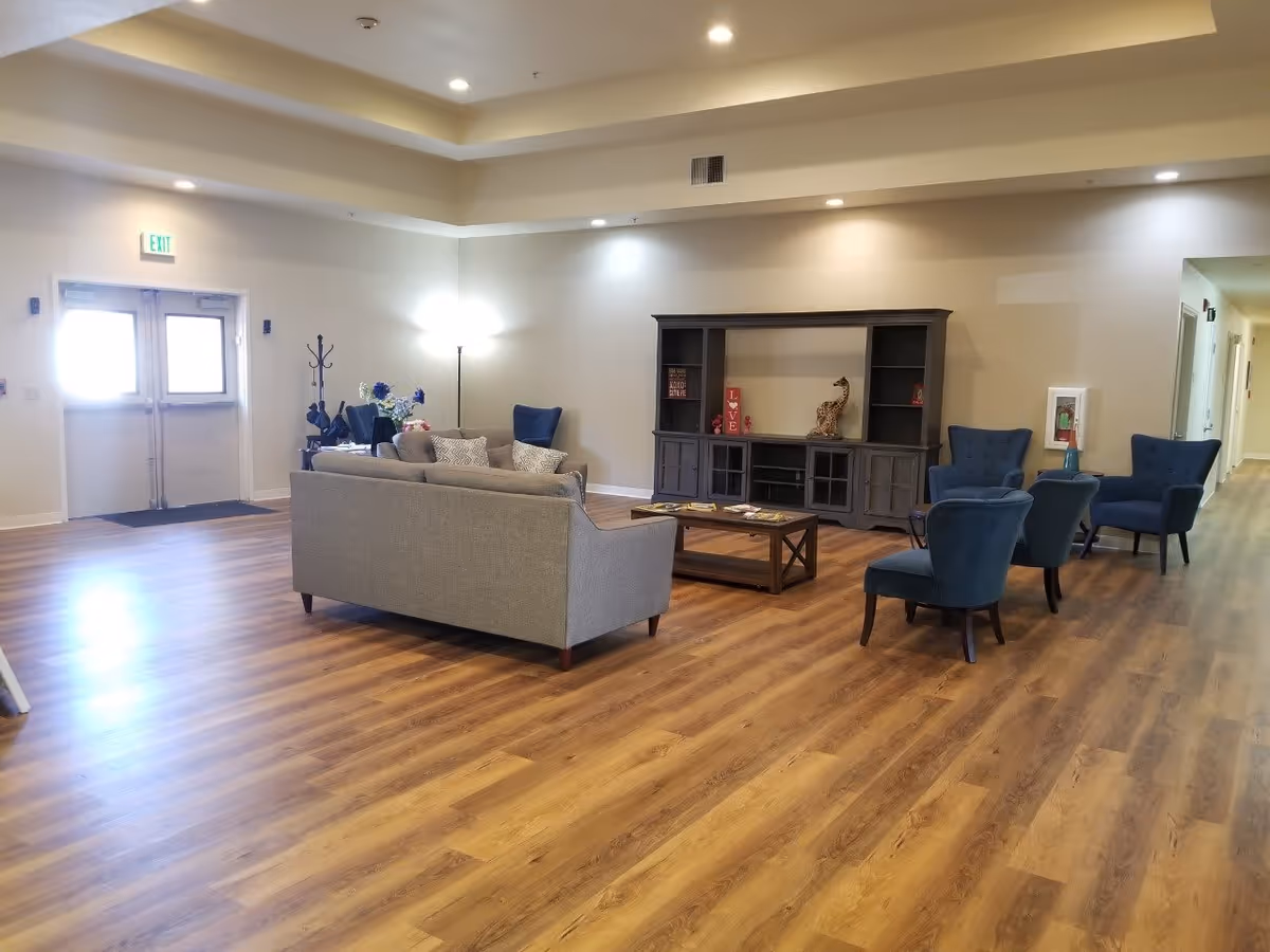 A spacious living room area with wooden flooring, a gray sofa with cushions, four blue armchairs, a wooden coffee table, and a large dark wooden entertainment center against the wall. There is a coat rack with some items hanging, a floor lamp, and a double door exit with an illuminated exit sign above it. The room has recessed lighting and a hallway leading to other rooms.