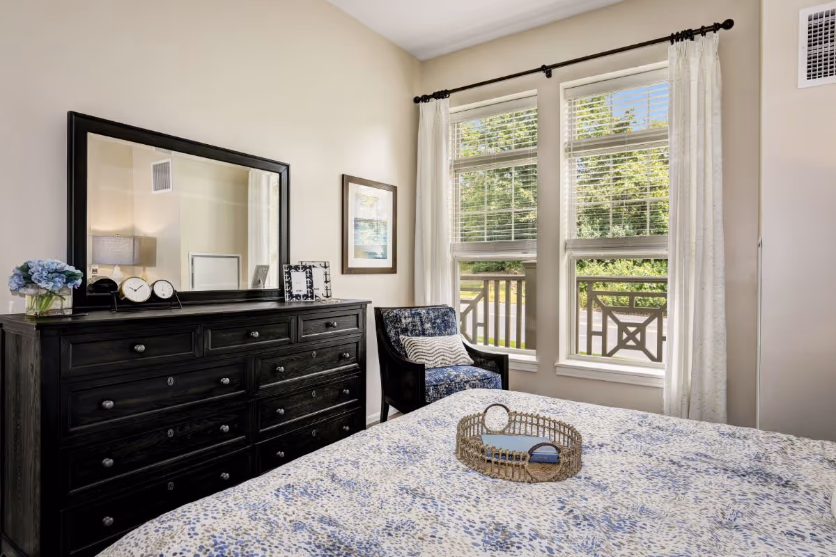 Sunlit bedroom with a patterned bedspread, a dark wood dresser topped with a large mirror and decorative items, and a blue upholstered chair by two windows overlooking greenery.