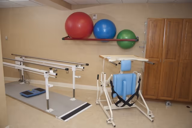 Physical therapy room with parallel bars for walking exercises, a blue supportive therapy chair with straps, a wooden cabinet, and three large exercise balls in red, blue, and green mounted on a wall rack.