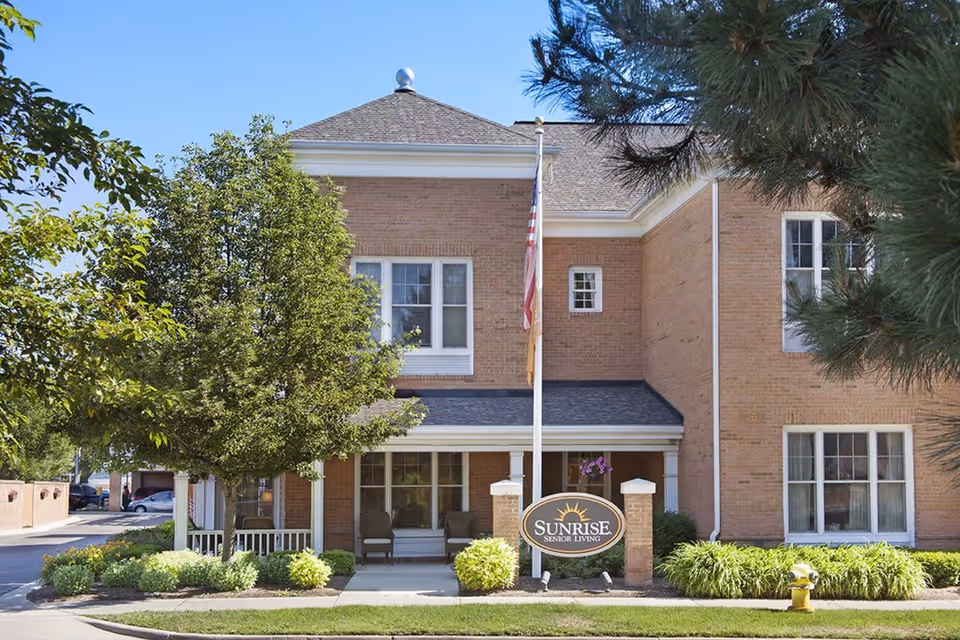 Exterior view of a two-story brick building with white trim, a small porch with chairs, an American flag on a flagpole, and a sign that reads 'Sunrise Senior Living' in front. There are trees and shrubs around the building under a clear blue sky.