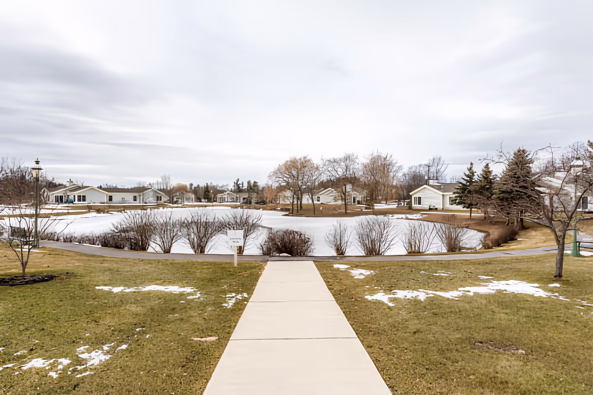 A paved walkway leads through a grassy area with patches of snow towards a frozen pond surrounded by leafless bushes and trees. Several single-story buildings are visible in the background under a cloudy sky.