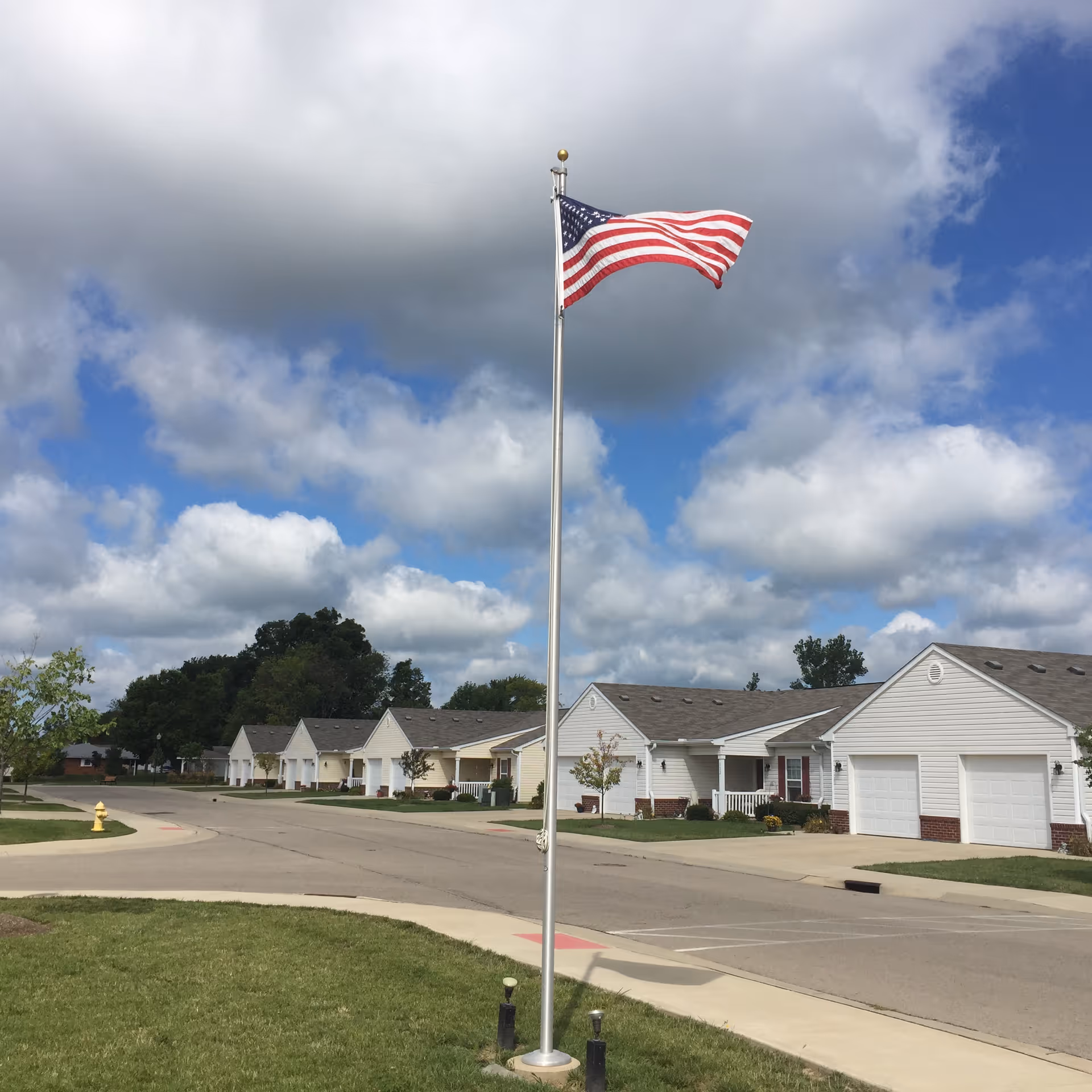 A row of single-story residential buildings with garages along a curved street under a partly cloudy sky. In the foreground, an American flag flies on a flagpole surrounded by grass and a sidewalk.
