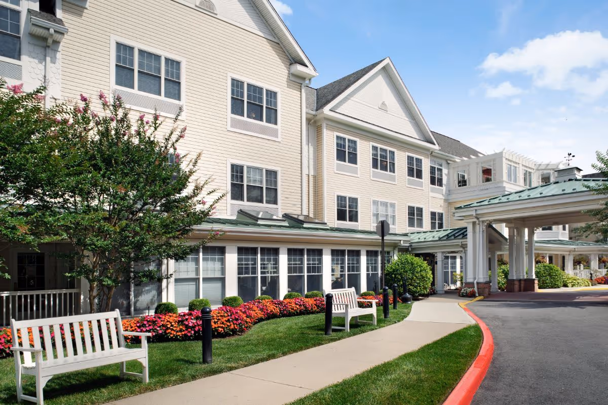 Front exterior of a multi-story senior living building with landscaped flowerbeds, benches, a walkway, and a covered entrance.