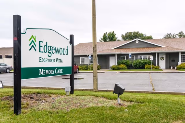 Front exterior of a single-story memory care facility with a large 'Edgewood' sign on a grassy lawn and a parking area behind it.