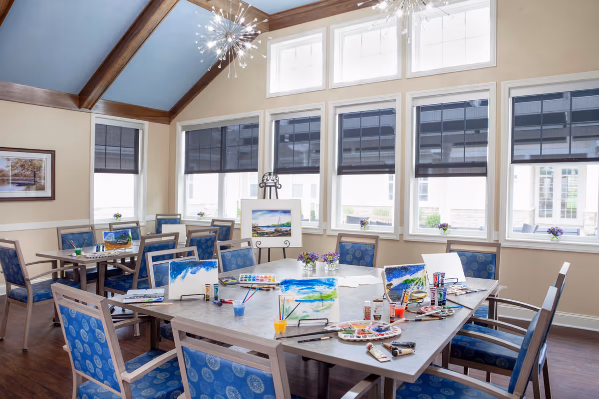 A bright and spacious activity room with a large table surrounded by blue cushioned chairs. The table is set up for painting with canvases, paintbrushes, paint tubes, palettes, and cups of water. The room has large windows with black roller shades, letting in natural light. The ceiling is vaulted with wooden beams and modern light fixtures. Small flower arrangements are placed on the windowsills and table.