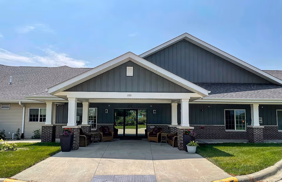 Front entrance of a single-story senior living building with a covered porte-cochere, outdoor seating, and landscaped lawn.
