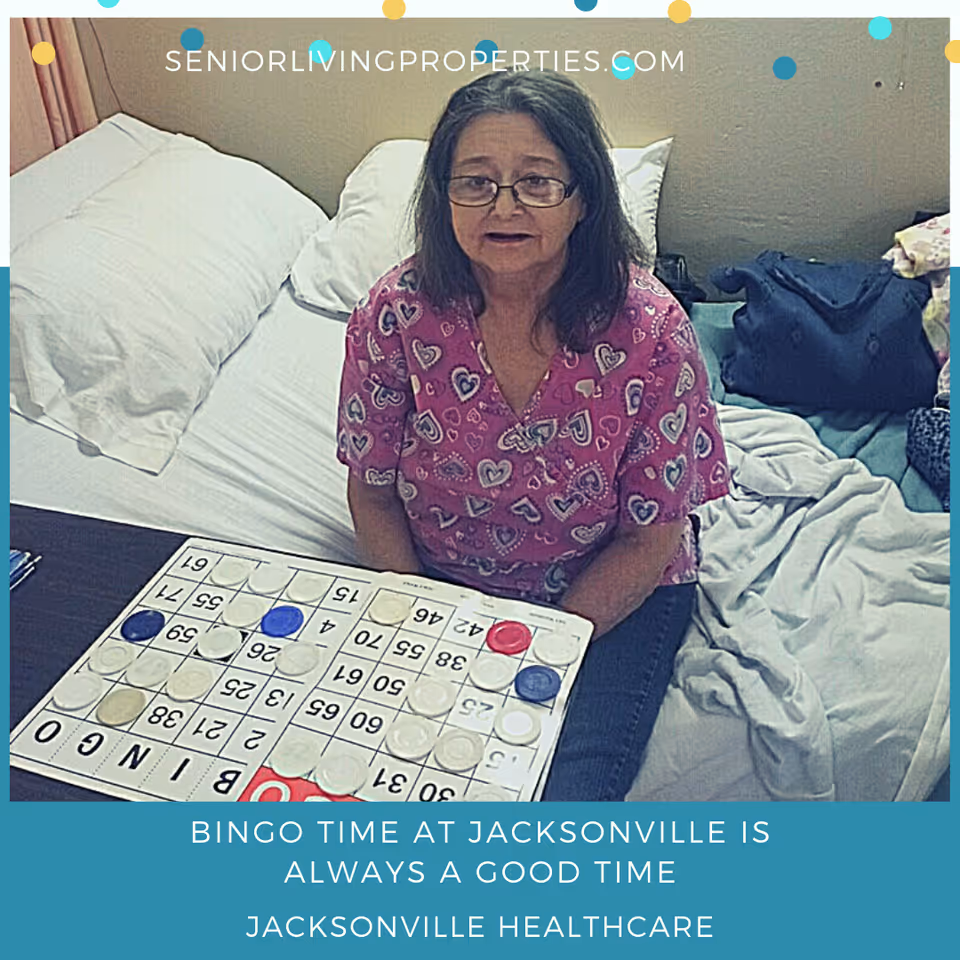 An elderly woman wearing glasses and a pink shirt with heart patterns is sitting on a bed playing bingo. The bed has white pillows and a light-colored blanket. There are some personal items and a blue bag on the bed beside her.