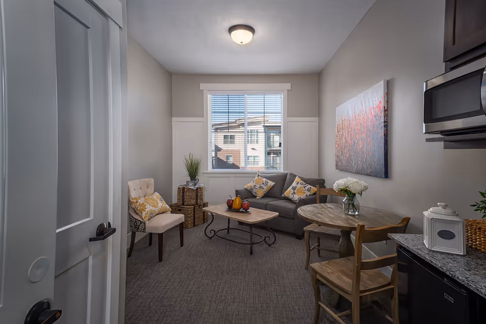 Furnished small living area with a sofa, chairs, coffee table and round dining table beside a kitchenette under a window.