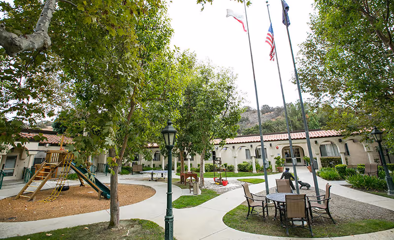 Outdoor courtyard area at Silverado Calabasas Memory Care Community featuring a playground with slide and climbing ropes, several trees, a round table with chairs, flagpoles with flags, and a single-story building with arched windows and a tiled roof in the background.