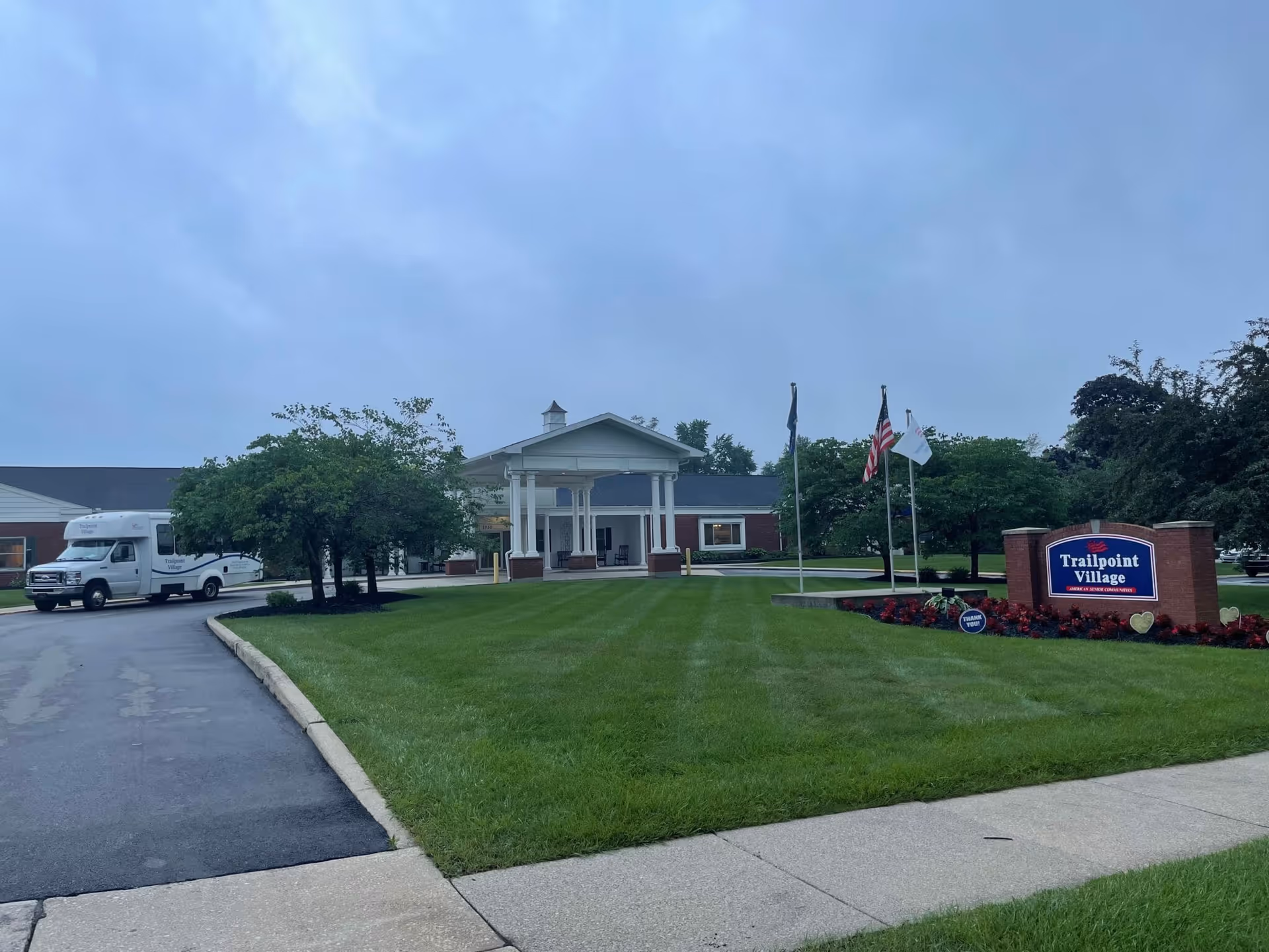 Exterior view of Trailpoint Village senior living facility showing a well-maintained lawn, a driveway with a parked shuttle bus, a building entrance with white columns, and a sign with the facility name surrounded by flowers and flags.