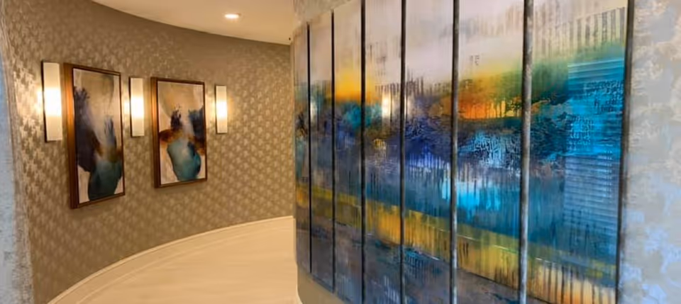 Curved interior hallway with large abstract panel art on the right, framed paintings and wall sconces on the left, and light wood flooring.