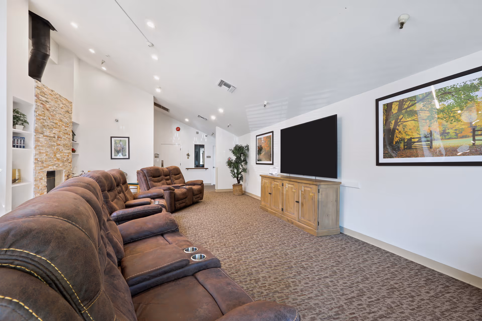 A spacious living room with brown leather reclining sofas facing a large flat-screen TV mounted on a wooden cabinet. The room has a high ceiling with recessed lighting, a stone fireplace on the left, framed nature artwork on the walls, and a potted plant near the back wall.