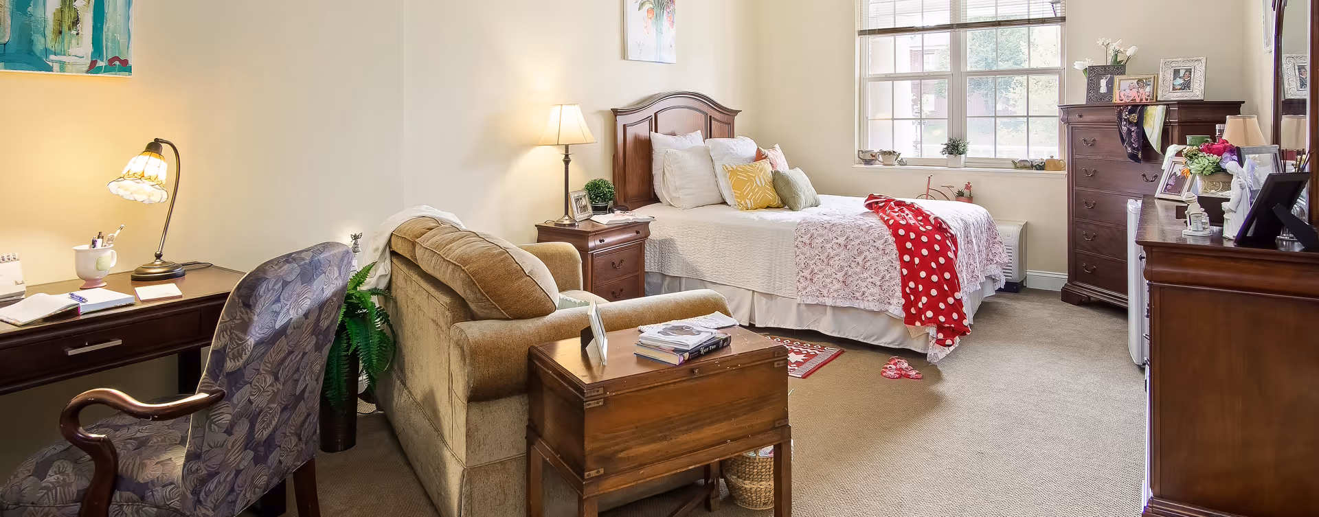 A cozy bedroom in a senior living facility featuring a bed with white and pink bedding and a red polka dot throw, a wooden nightstand with a lamp, a comfortable beige armchair, a wooden desk with a floral-patterned chair and a lamp, a wooden dresser, and a window letting in natural light.