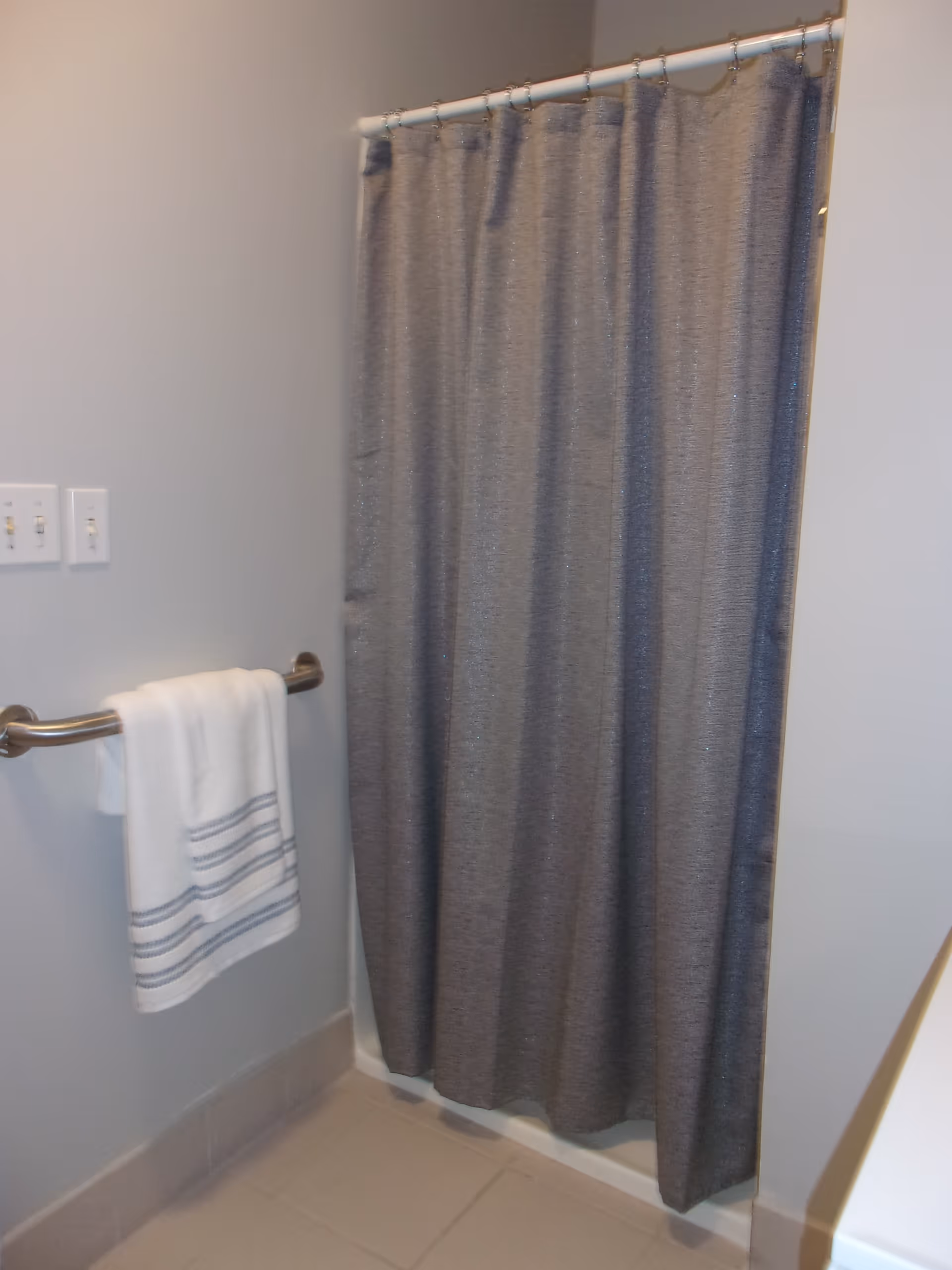 A bathroom corner featuring a gray shower curtain hanging from a white rod, a towel rack with a white towel that has gray stripes, and light gray tiled floor and walls.