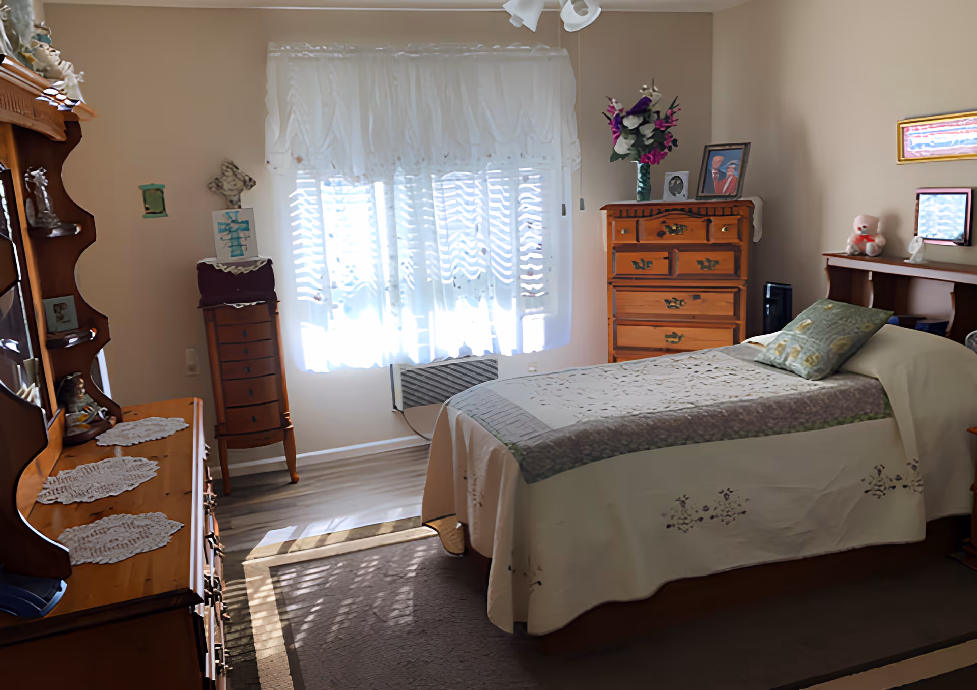 A sunlit bedroom with a neatly made twin bed, wooden dressers and lace-curtained window.