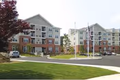 Front view of two multi-story residential buildings with landscaped lawn, a parking area, and flagpoles in front.