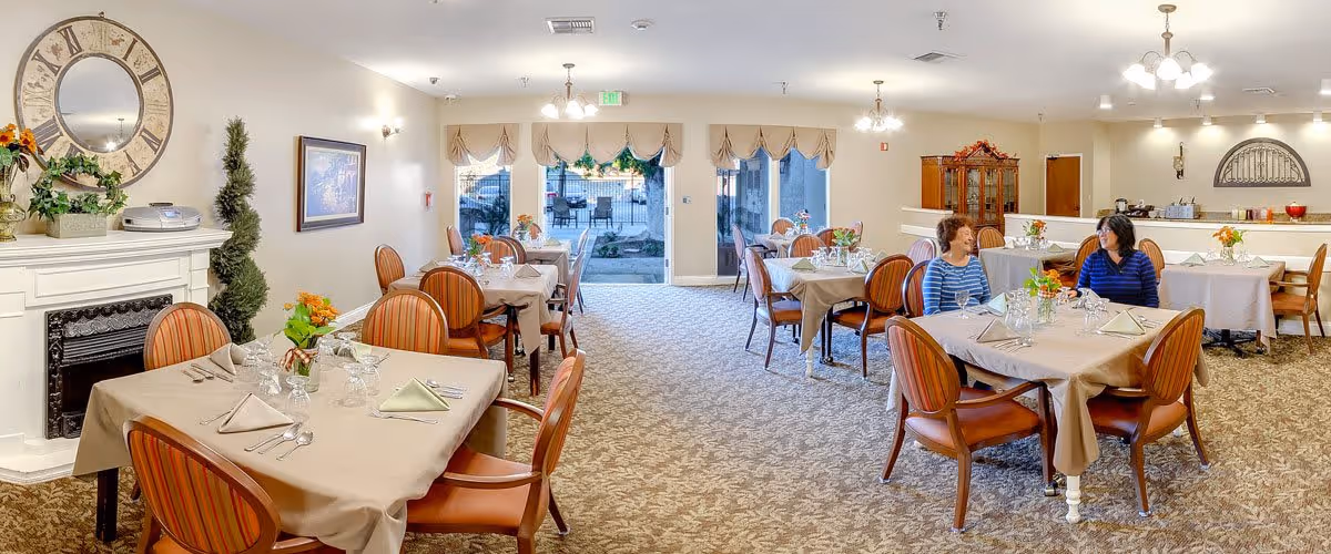A bright and spacious dining room in a senior living facility with multiple tables covered in beige tablecloths, each set with folded napkins, glassware, and silverware. Two women are seated and conversing at one of the tables. The room features carpeted flooring, a decorative fireplace with plants and a large clock above it, wall sconces, and large windows with valances letting in natural light. There is a china cabinet and a counter area in the background.