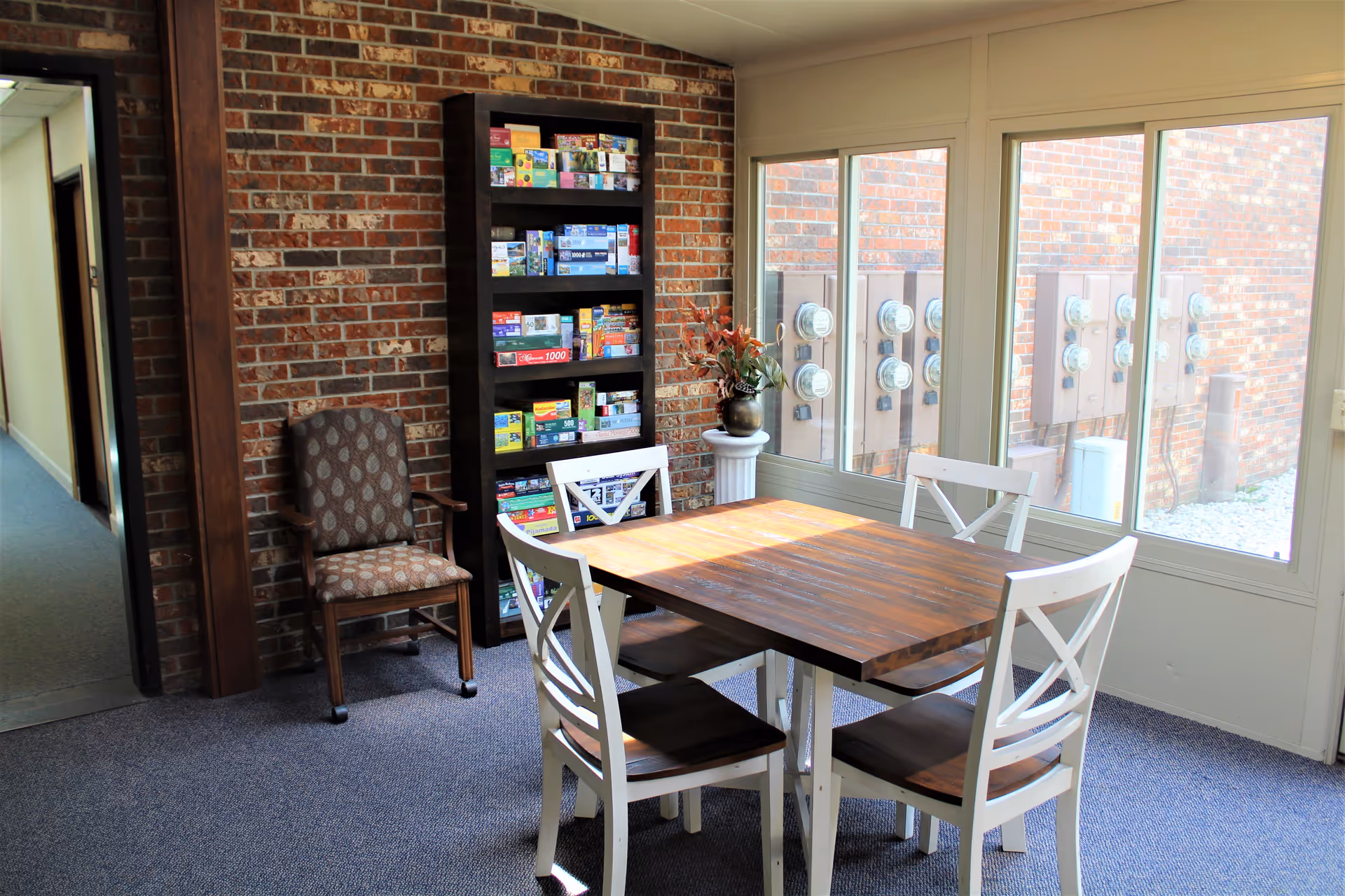 A bright room with a wooden table and four white chairs with wooden seats. There is a tall black bookshelf filled with puzzles and games against a brick wall. A patterned armchair is placed next to the bookshelf. Large windows show an exterior brick wall with multiple utility meters. A decorative plant in a vase is on a white pedestal near the window.