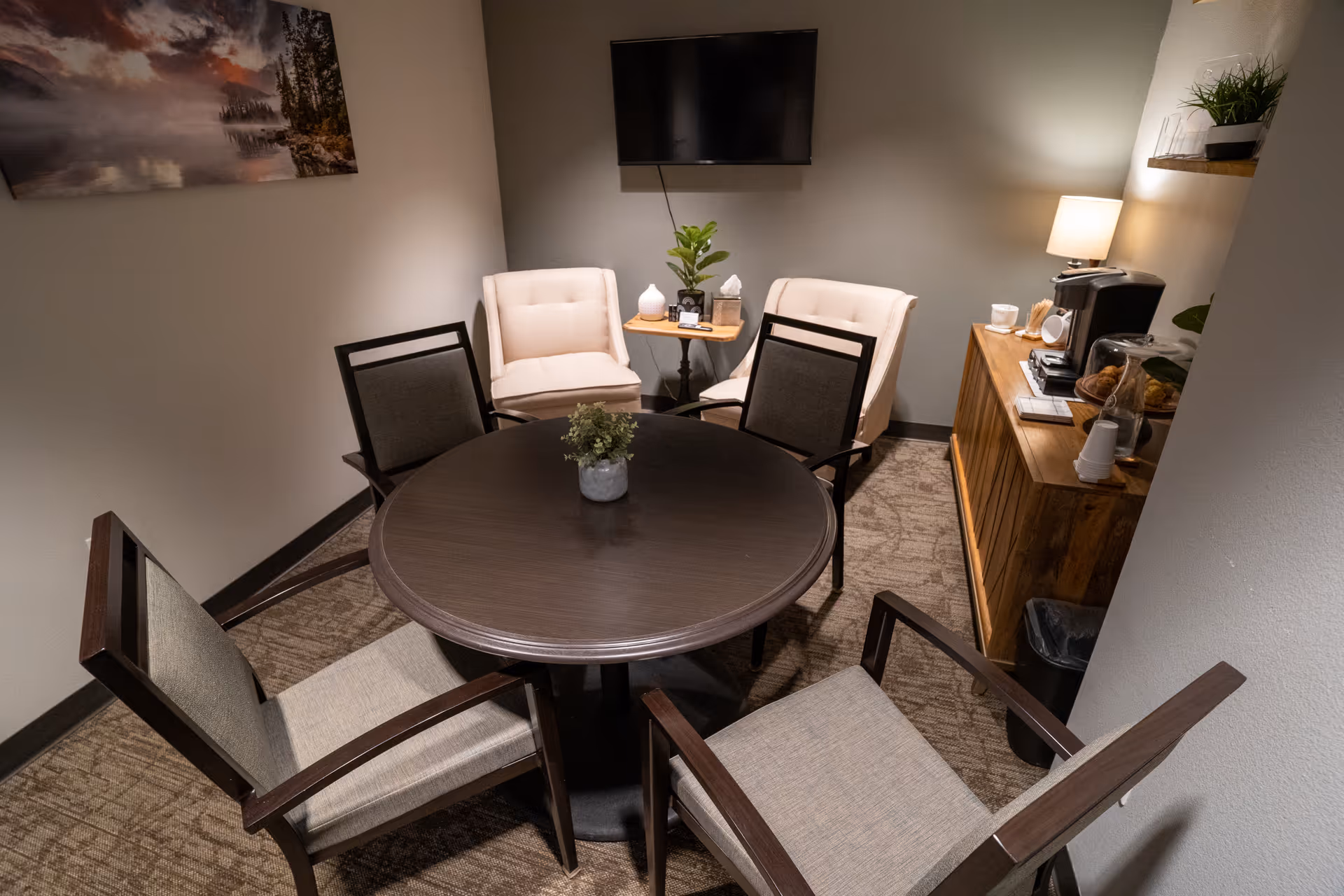 A small cozy seating area with a round dark wooden table surrounded by four cushioned chairs. Two beige armchairs are placed against the wall beneath a mounted flat-screen TV. A wooden sideboard holds a lamp, coffee maker, cups, and snacks. A small plant is on the table and another on a side table between the armchairs. A landscape painting hangs on the wall.