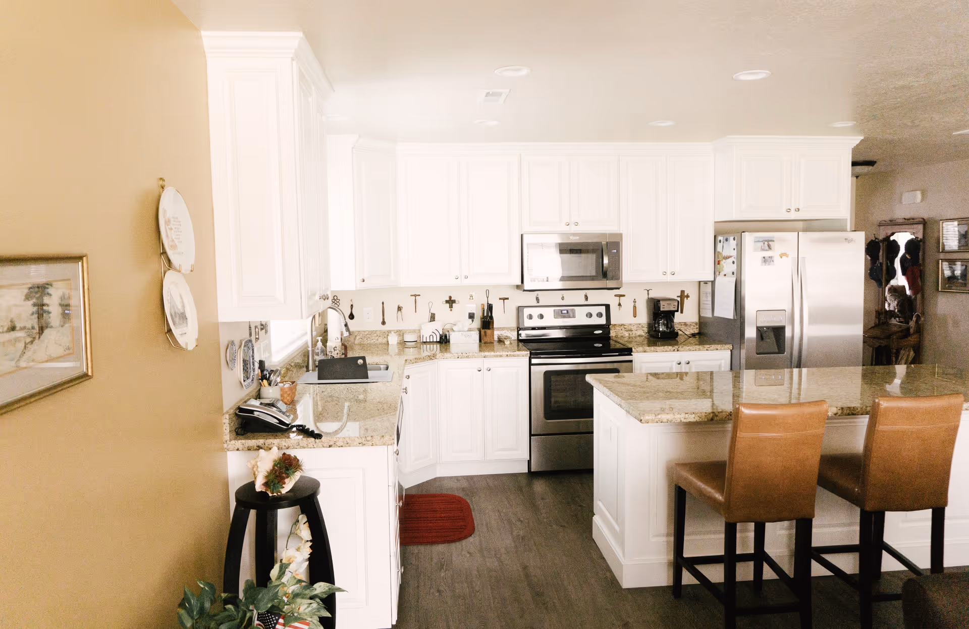 A bright kitchen with white cabinets, granite countertops, and stainless steel appliances including a stove, microwave, and refrigerator. There is a kitchen island with two brown leather bar stools. The floor is wood, and there are decorative items on the walls and counters.