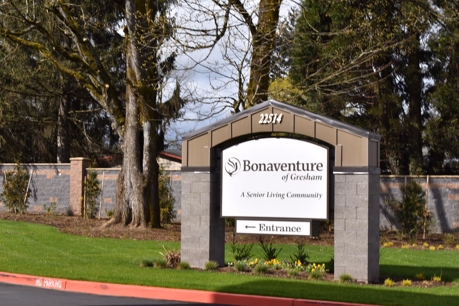 Outdoor view of a sign for Bonaventure of Gresham, a senior living community, with the address number 22514 displayed above. The sign is set on a landscaped area with grass, flowers, and trees in the background.