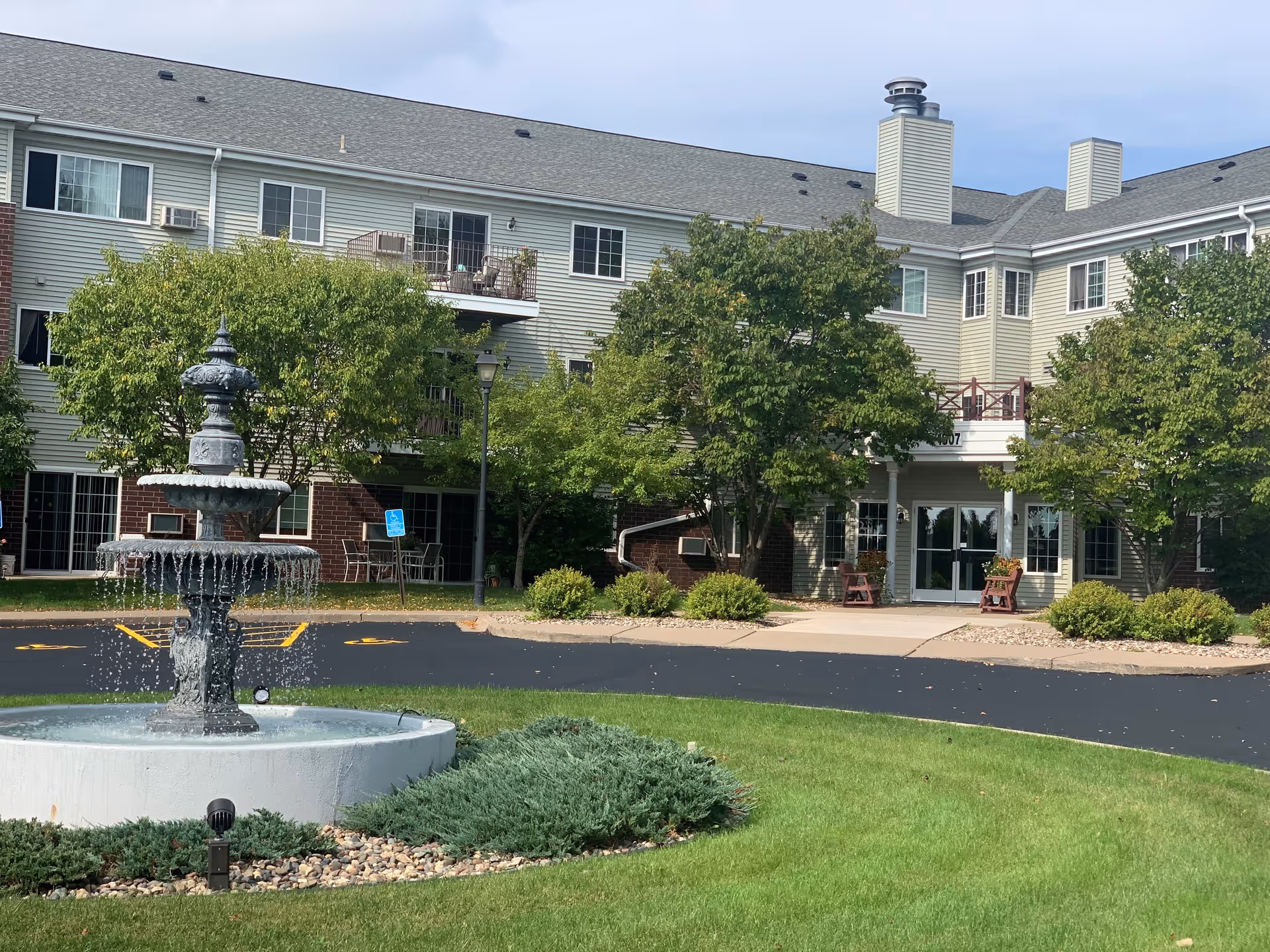 Exterior view of Norgard Court Apartments, showing a three-story building with balconies, several trees, a fountain in the foreground, and a paved driveway leading to the entrance.