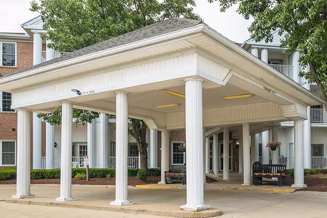 Covered entrance area of a senior living facility with white columns and benches, surrounded by trees and a multi-story brick and white building in the background.