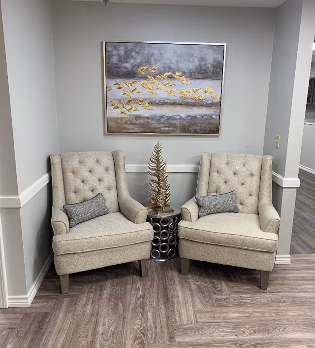 Two beige tufted armchairs with gray rectangular pillows are positioned side by side in a corner with light gray walls. Between the chairs is a small round metallic side table with a decorative gold leaf sculpture on top. Above the chairs hangs a large framed abstract painting featuring gold birds in flight against a gray and white background. The floor is covered with wood-patterned vinyl planks.