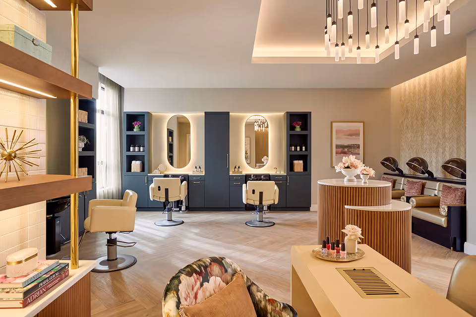 A modern and elegant salon area with two styling chairs in front of illuminated oval mirrors, shelving units with decorative items, and a seating area with hair drying stations. The room features warm lighting, a chandelier, and a table with nail polish and flowers.