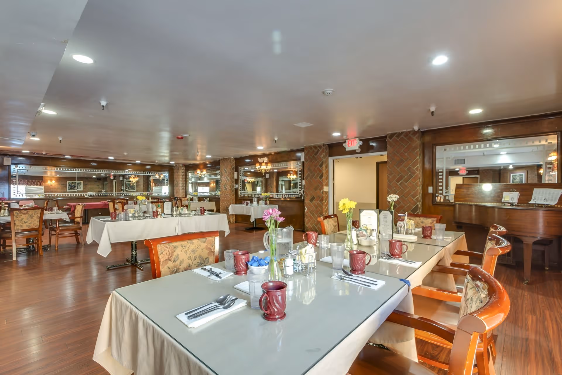 A spacious dining room in a senior living facility with several tables covered in white tablecloths and set with red mugs, silverware, and glassware. The room features wooden chairs with floral upholstery, a polished wooden floor, large mirrors on the walls, and a piano in the corner. Fresh flowers in vases decorate the tables, and warm lighting creates a welcoming atmosphere.