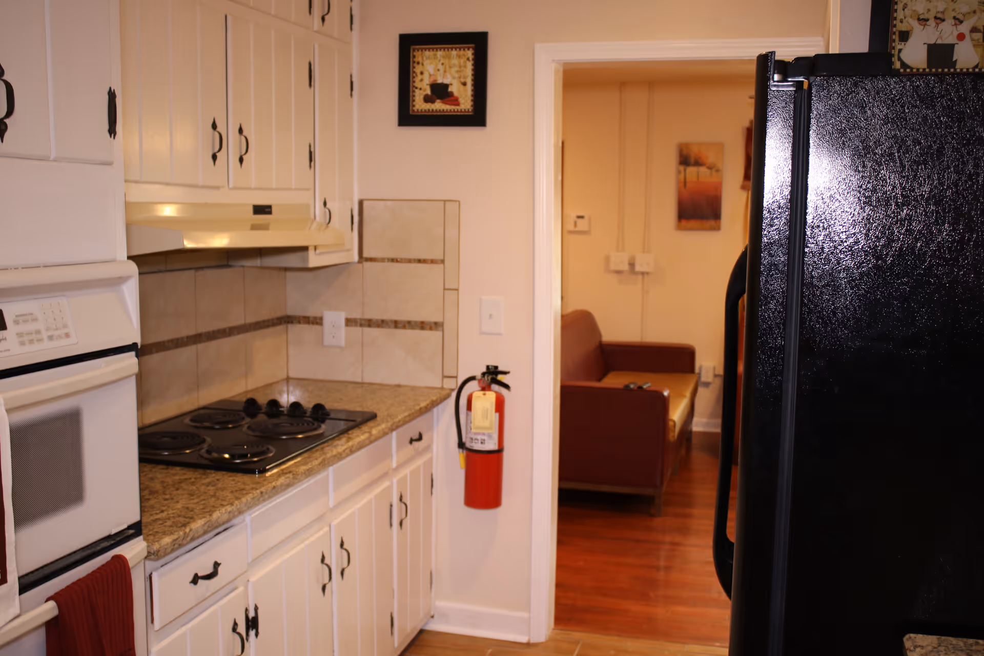A kitchen area with white cabinets, a granite countertop, an electric stovetop, and a built-in oven. A black refrigerator is visible on the right side. A fire extinguisher is mounted on the wall near the doorway, which leads to a room with a brown couch and wall art.