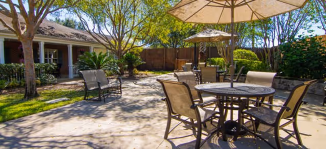 Sunny outdoor courtyard with round patio tables and umbrellas, seating, trees, and a building facade in the background.