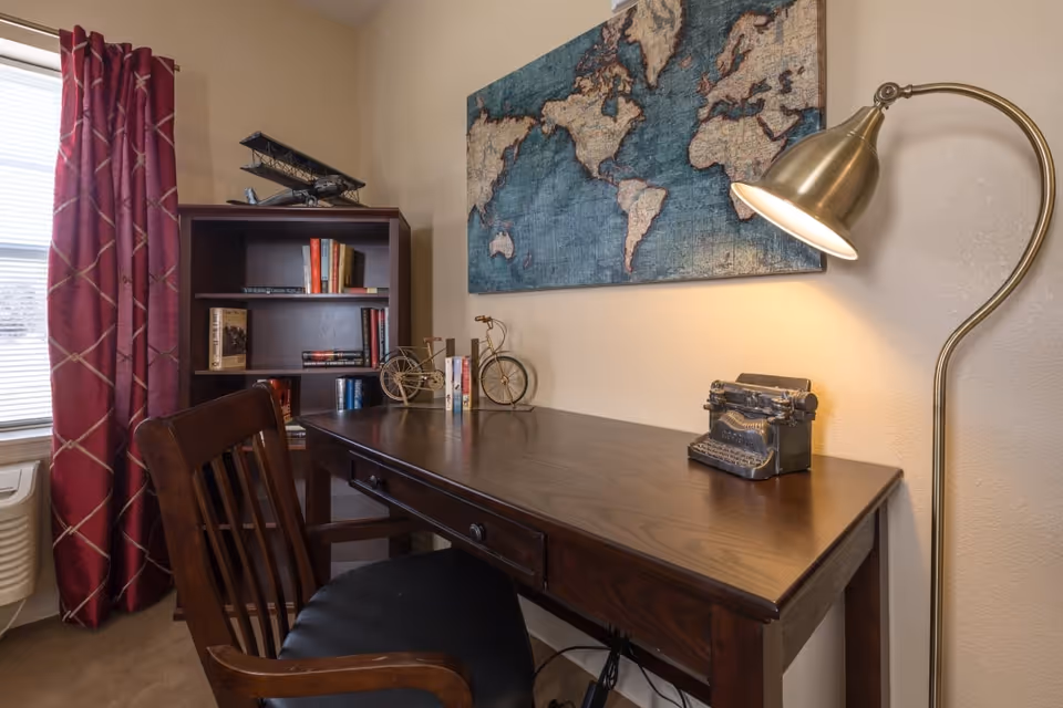 A cozy study area with a dark wooden desk and matching chair. On the desk are decorative items including a vintage typewriter, a small metal bicycle bookend holding books, and a brass floor lamp providing light. Behind the desk is a bookshelf with various books and a model airplane on top. A large world map artwork hangs on the wall above the desk. A window with red curtains is visible to the left.