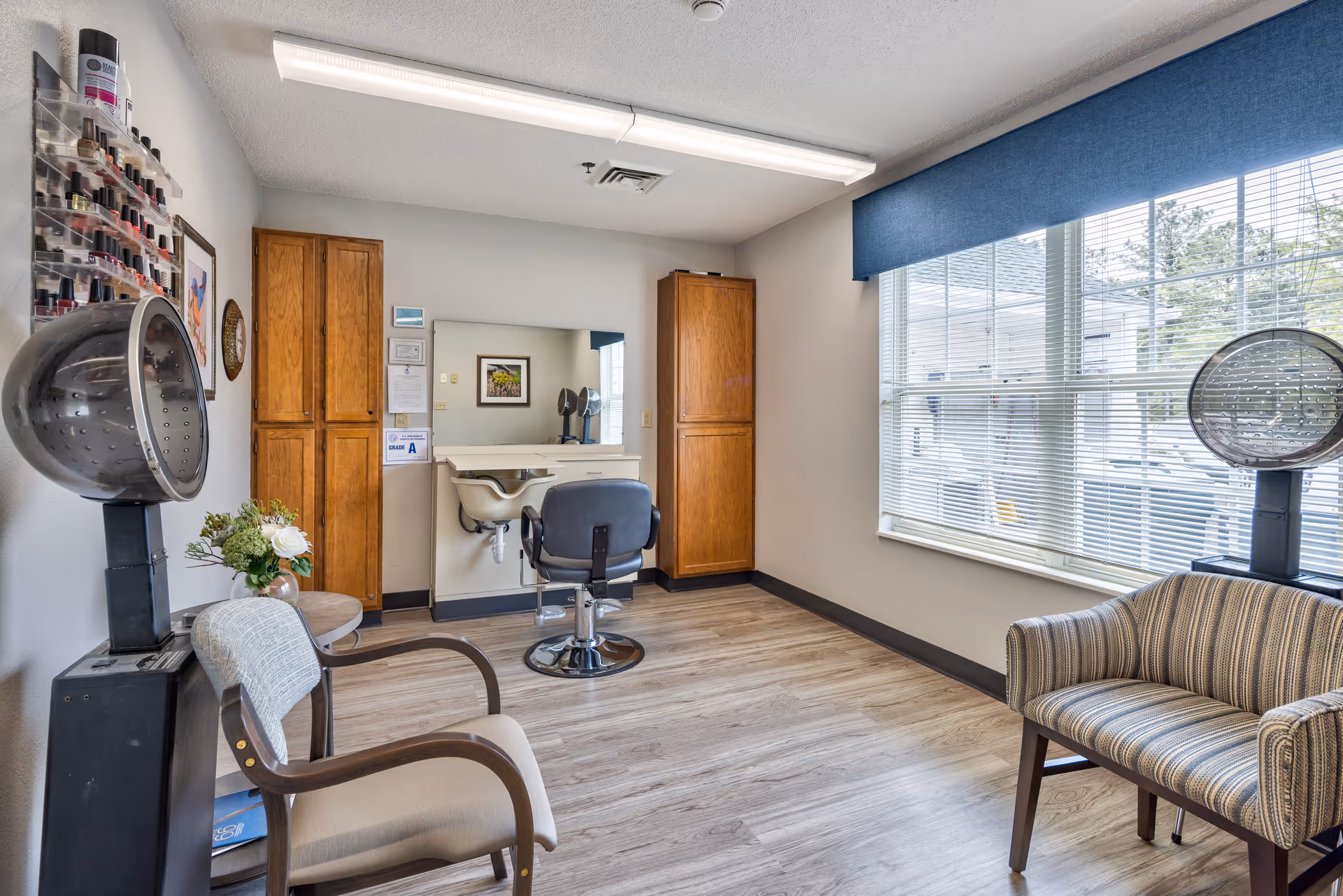 A bright salon room with two hair drying chairs, a styling chair in front of a mirror, wooden cabinets, a small table with a flower vase, and a large window with blue blinds letting in natural light.