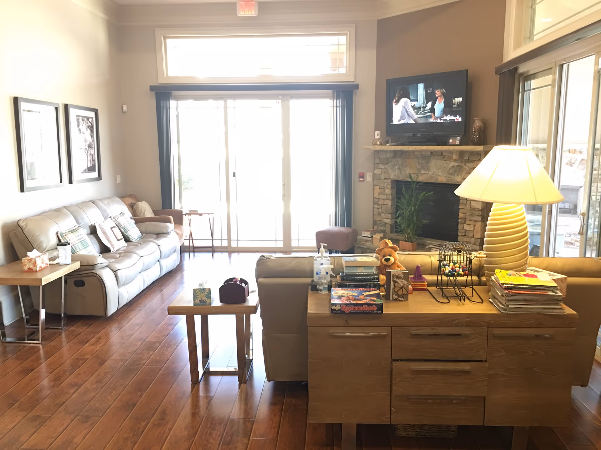A bright living room with wooden flooring featuring a beige leather sofa with pillows, a wooden side table with tissues and a cup, and a wooden console table with board games, books, a teddy bear, a lamp, and a hand sanitizer bottle. In the background, there is a stone fireplace with a TV mounted above it, a potted plant, and large sliding glass doors letting in natural light.