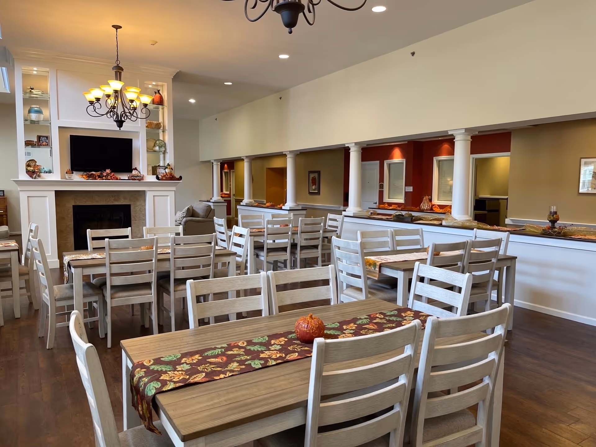 A spacious dining room with multiple wooden tables and chairs arranged neatly. Each table has a decorative runner with autumn leaves and a small pumpkin centerpiece. The room features a fireplace with a mounted TV above it, flanked by built-in shelves displaying decorative items. There is a chandelier hanging from the ceiling and white columns separating the dining area from an adjacent space with red accent walls.