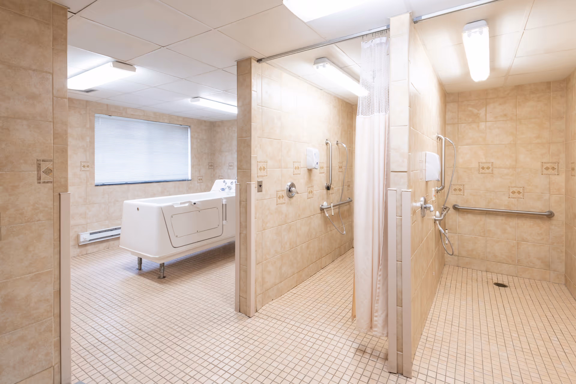 A spacious tiled bathroom area with two open shower stalls equipped with grab bars and handheld showerheads, and a large walk-in bathtub near a window with blinds. The walls and floor are covered with beige tiles, and the ceiling has fluorescent lighting.