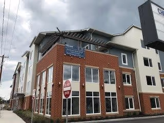 Exterior view of a multi-story senior living facility building with a combination of red brick and light-colored siding under a cloudy sky. A stop sign is visible in front of the building.