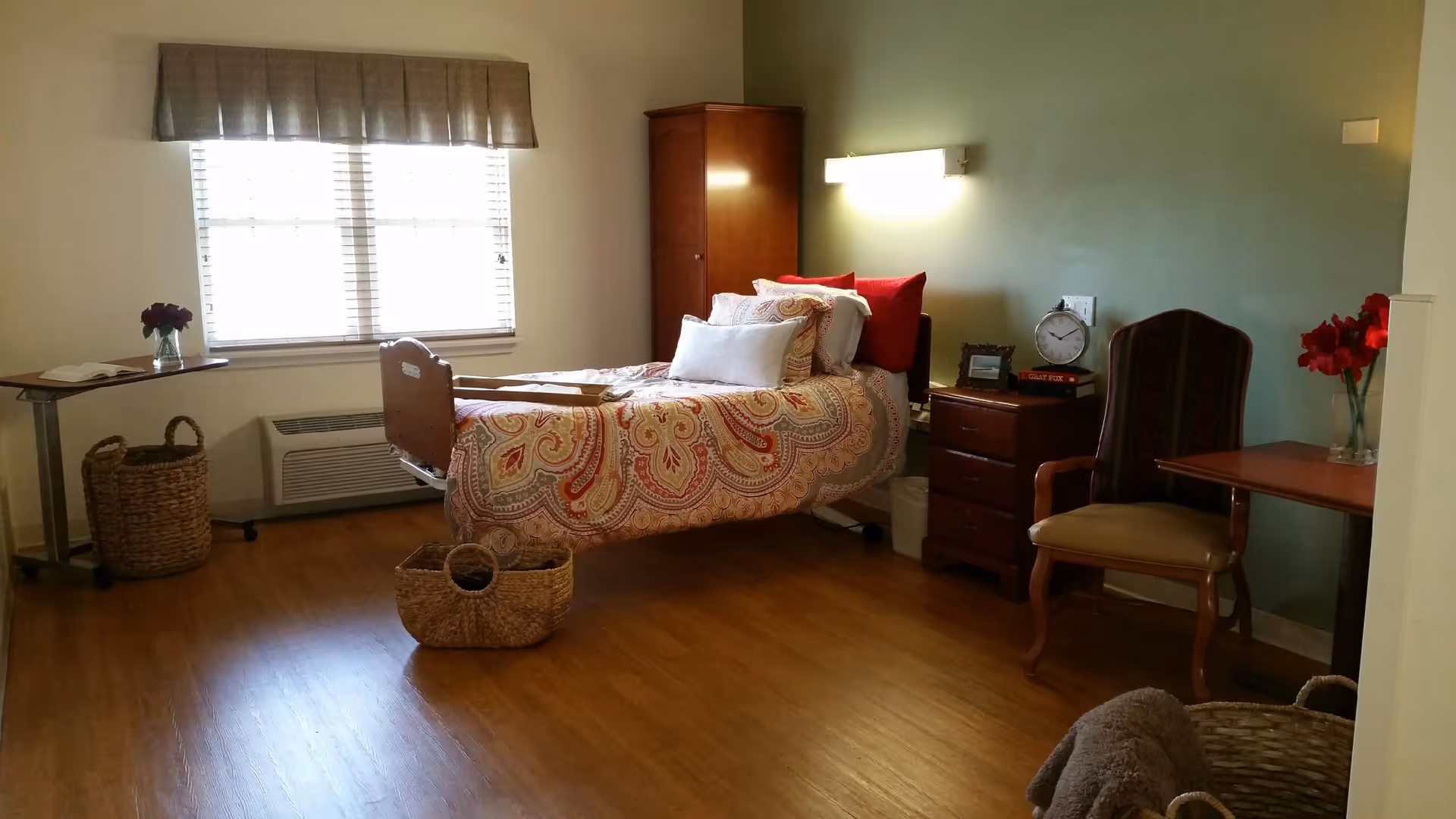 A neatly arranged bedroom in a senior living facility with a single bed featuring a colorful paisley-patterned bedspread and multiple pillows. The room has wooden flooring, a window with blinds and a valance, a wooden wardrobe, a bedside table with a clock and books, a chair, a small table with a vase of red flowers, and woven baskets on the floor.