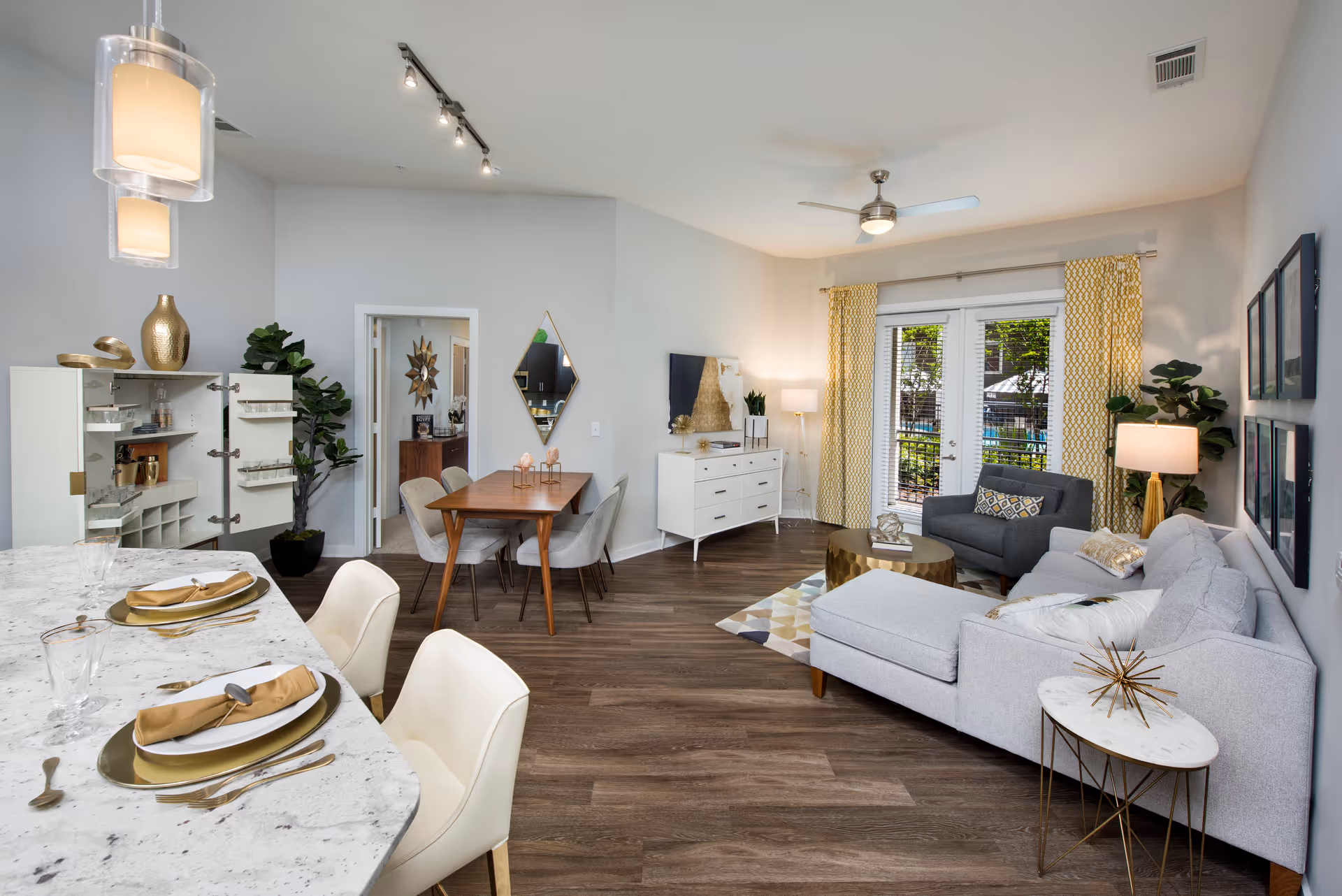 Open-plan living and dining area with a light-gray sectional chaise, armchair, wooden dining table, and a marble-topped kitchen island set for dining.