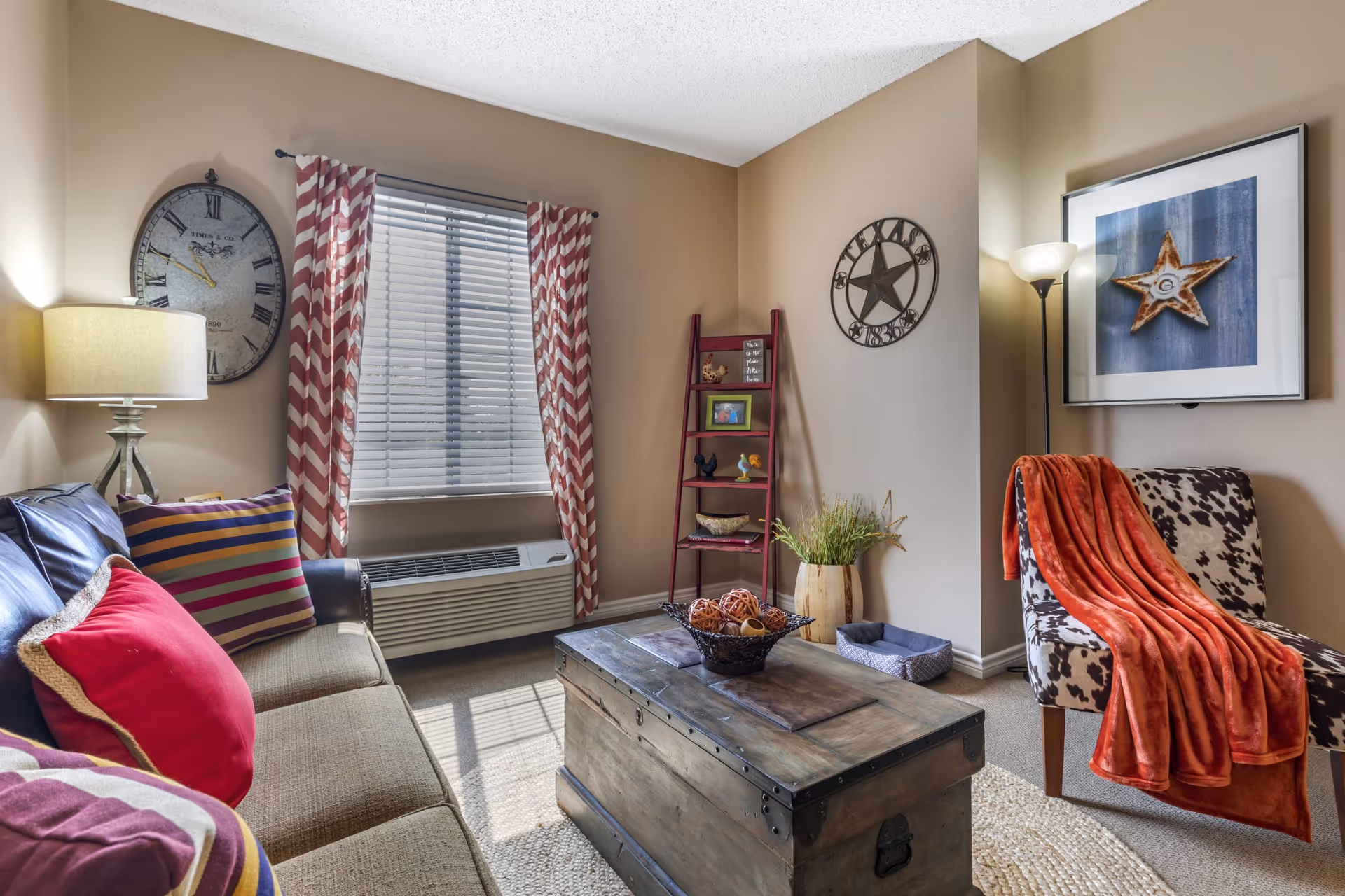 Cozy living room with a sofa with colorful pillows, a wooden trunk coffee table, a patterned armchair draped with an orange throw, and wall decor.