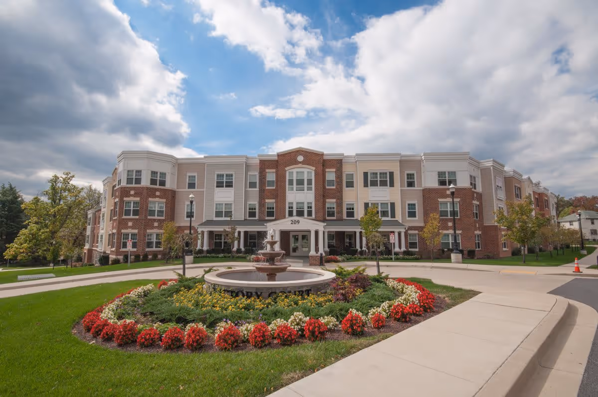 Front exterior view of Victory Court Senior Apartments, a three-story building with a mix of brick and light-colored siding. In front of the building is a circular driveway with a landscaped roundabout featuring a fountain and colorful flowers. The sky is partly cloudy.