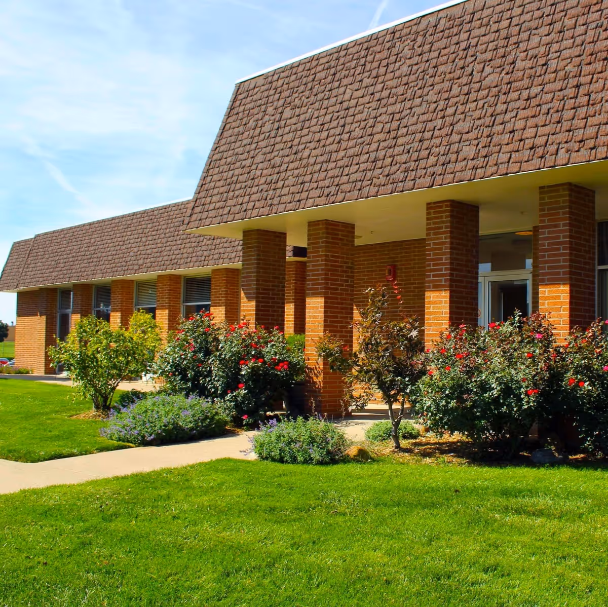 Exterior view of a brick building with a brown shingled roof, surrounded by green grass and landscaped bushes with red flowers under a clear blue sky.