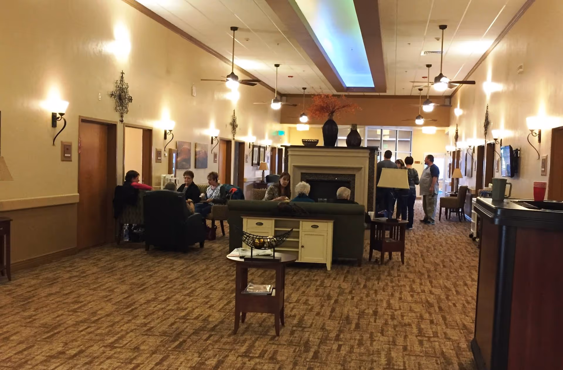 A spacious senior living community common area with carpeted floors, beige walls, and ceiling fans. Several seniors are seated on sofas and chairs near a central fireplace, while a few people stand and converse in the background. The room is warmly lit with wall sconces and pendant lights, and decorated with vases and wall art.
